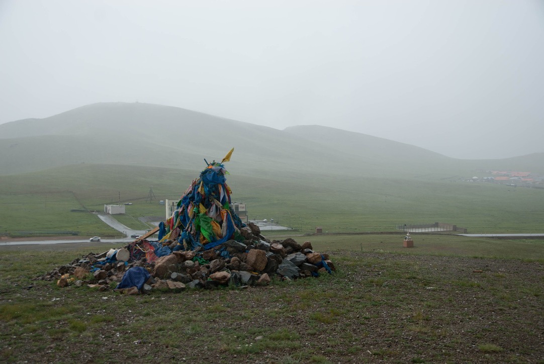 Stone ovoo with weathered blue prayer scarves on a rainy green pass.